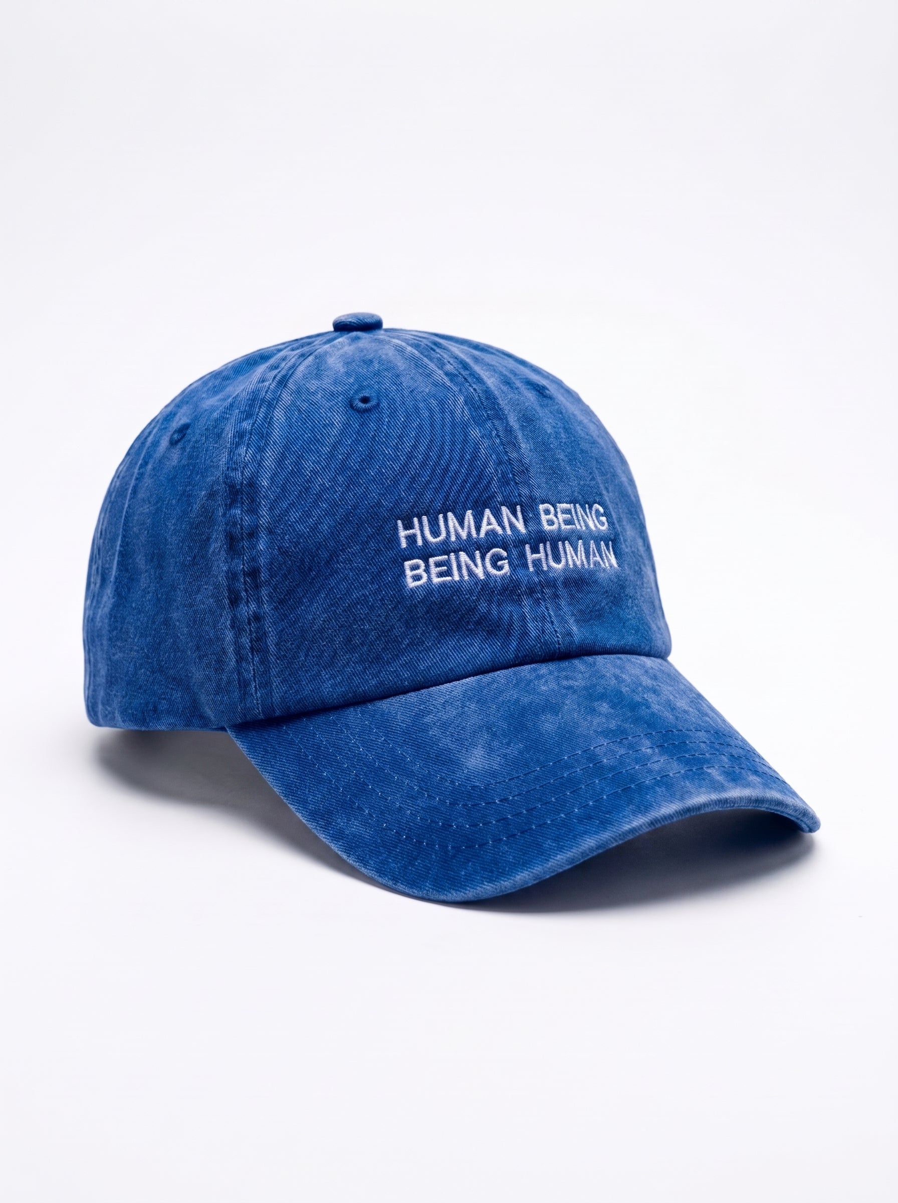 A blue washed cotton baseball cap with white embroidered text reading 'HUMAN BEING BEING HUMAN' on the front, shown on a clean white background.