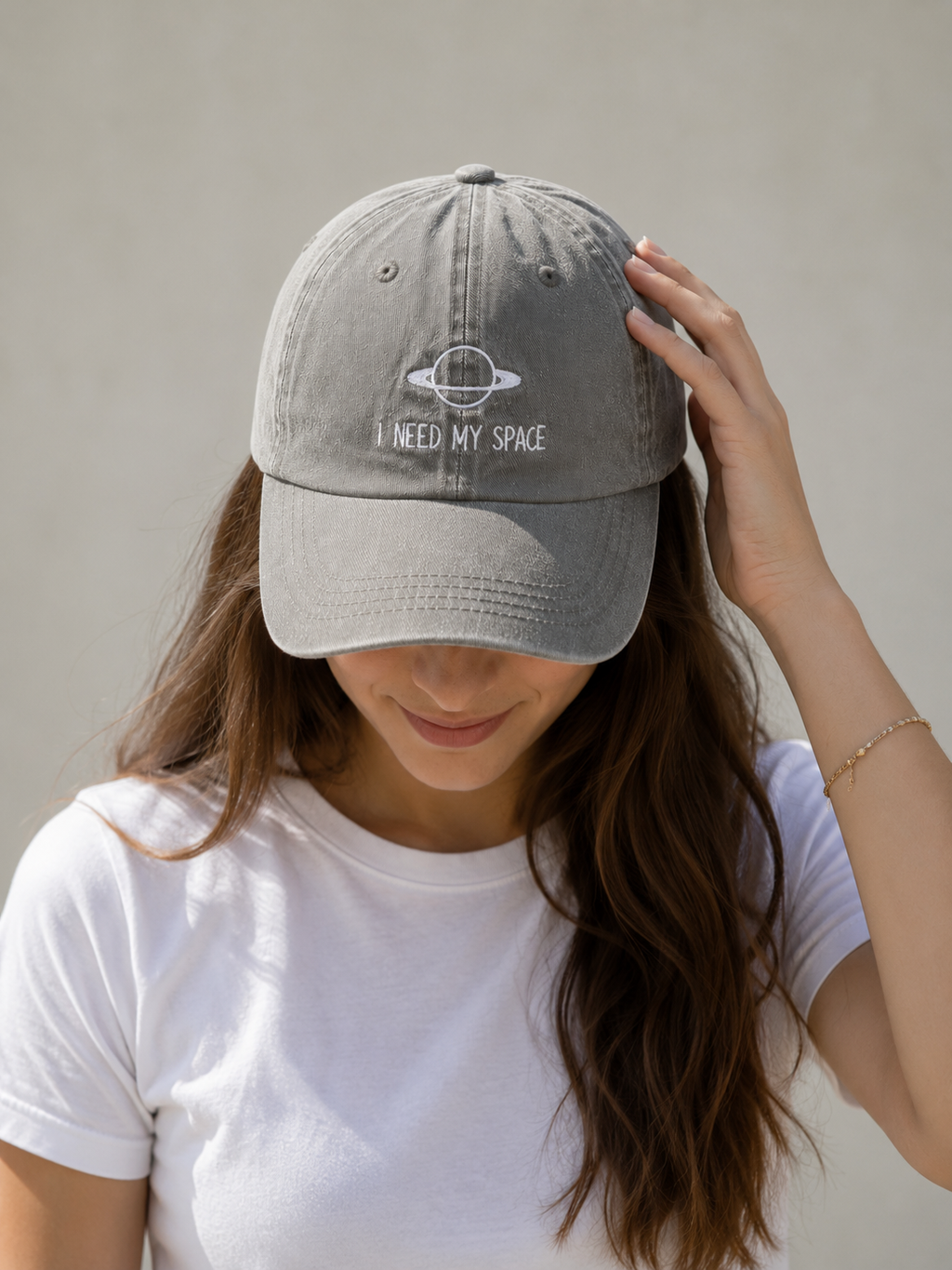 A woman wearing a light gray EMBROIDERED CAP with a white embroidered design of a ringed planet and the text 'I NEED MY SPACE'.