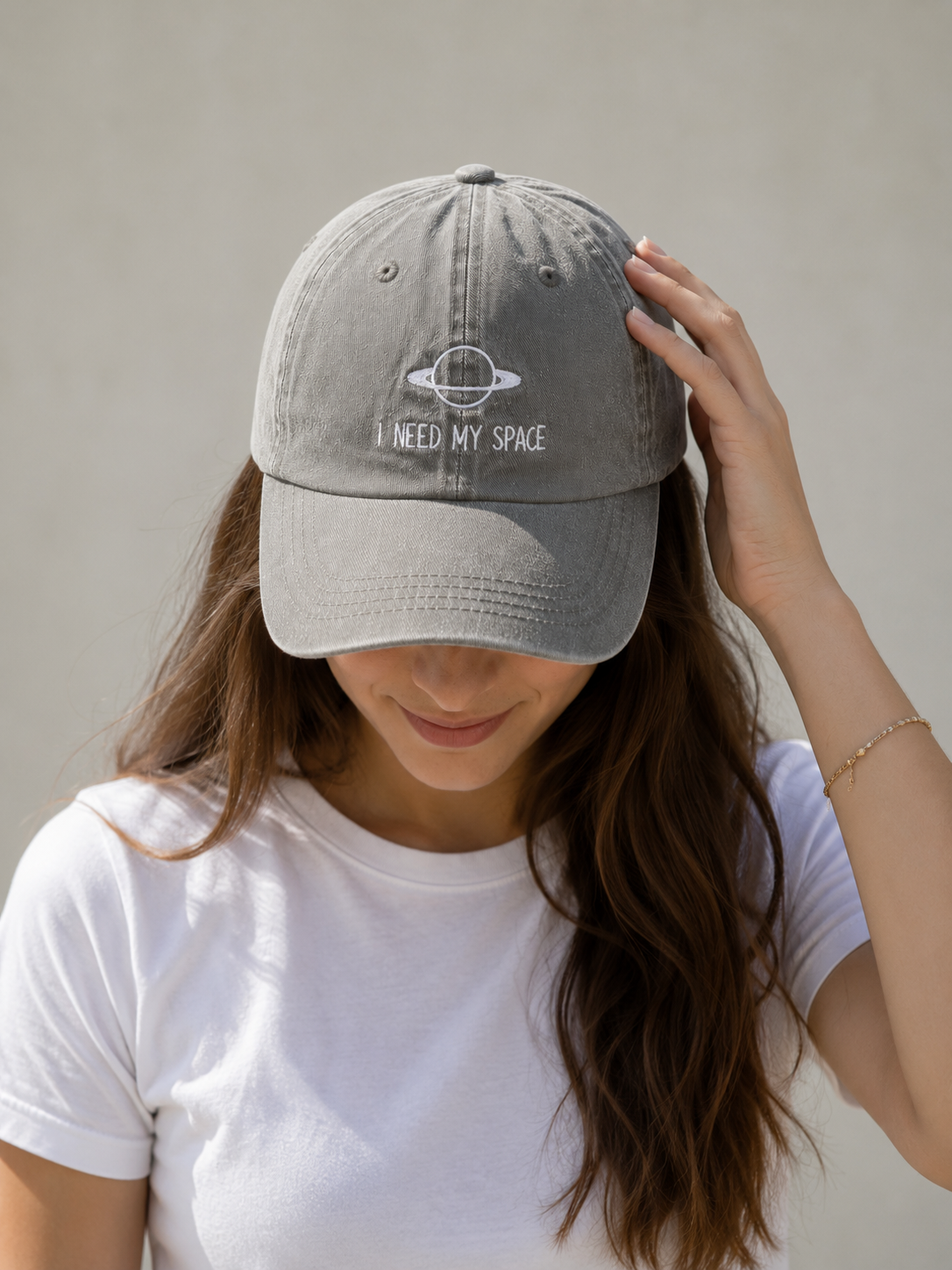 A woman wearing a light gray EMBROIDERED CAP with a white embroidered design of a ringed planet and the text 'I NEED MY SPACE'.