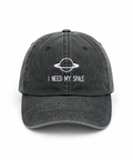 A dark gray washed baseball cap with a white embroidered graphic of a ringed planet and the text 'I NEED MY SPACE' on the front, shown against a plain white background.