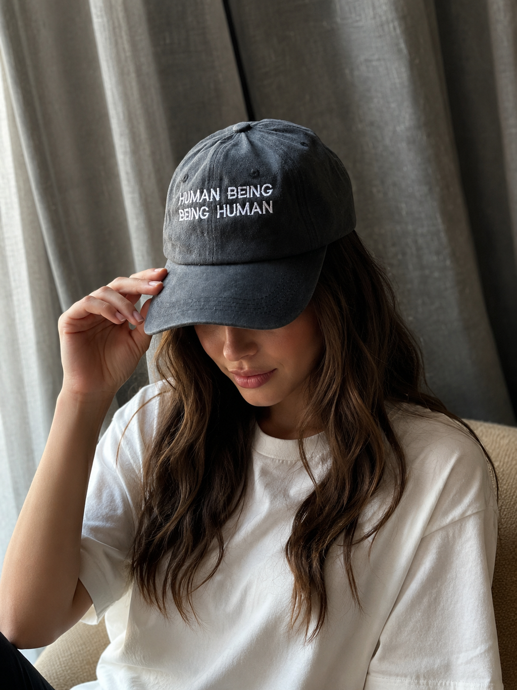 A woman wearing a dark gray washed baseball cap with white embroidered text reading 'HUMAN BEING BEING HUMAN' and a white t-shirt, posing indoors in front of gray curtains.