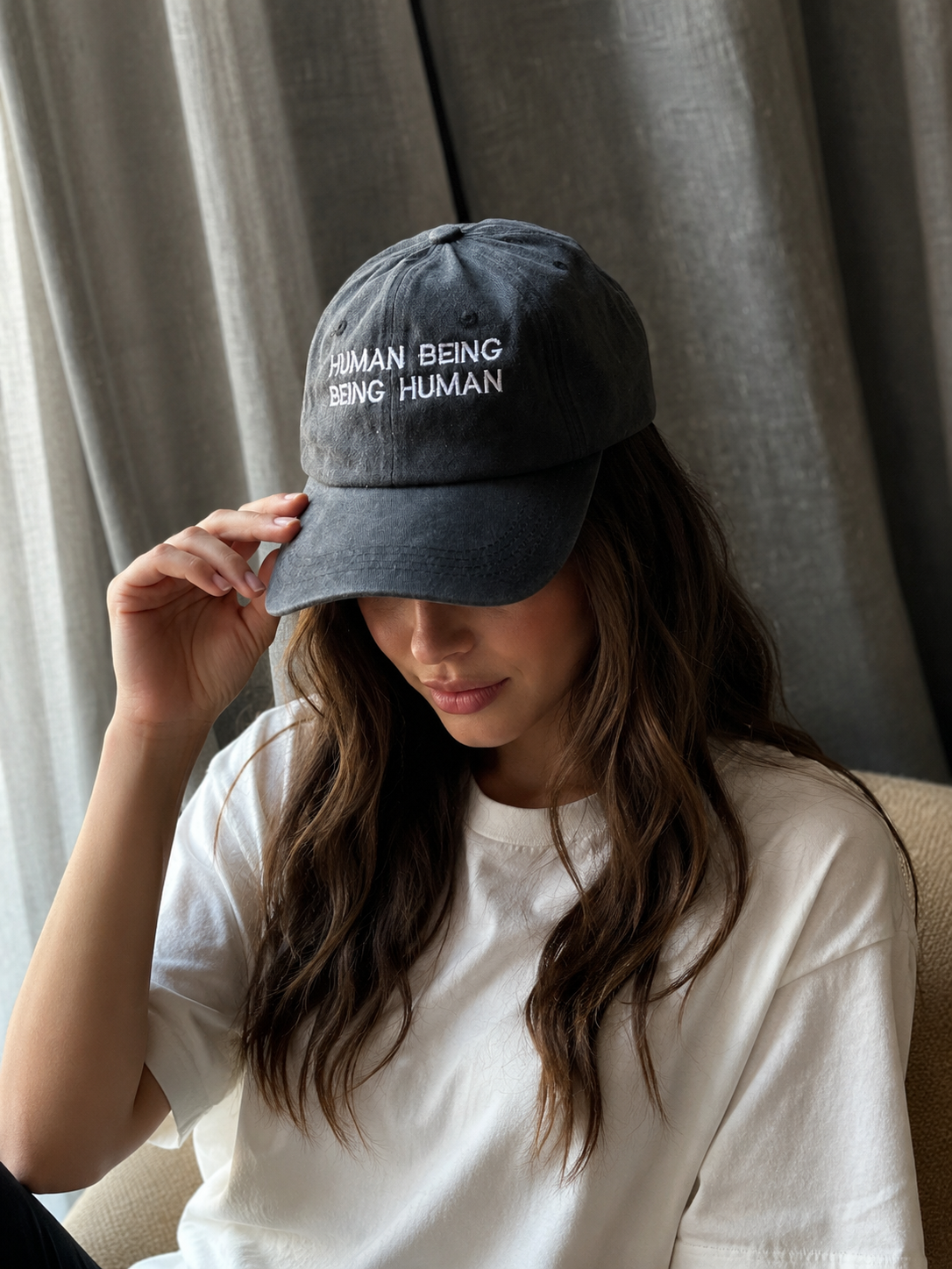 A woman wearing a dark gray washed baseball cap with white embroidered text reading 'HUMAN BEING BEING HUMAN' and a white t-shirt, posing indoors in front of gray curtains.