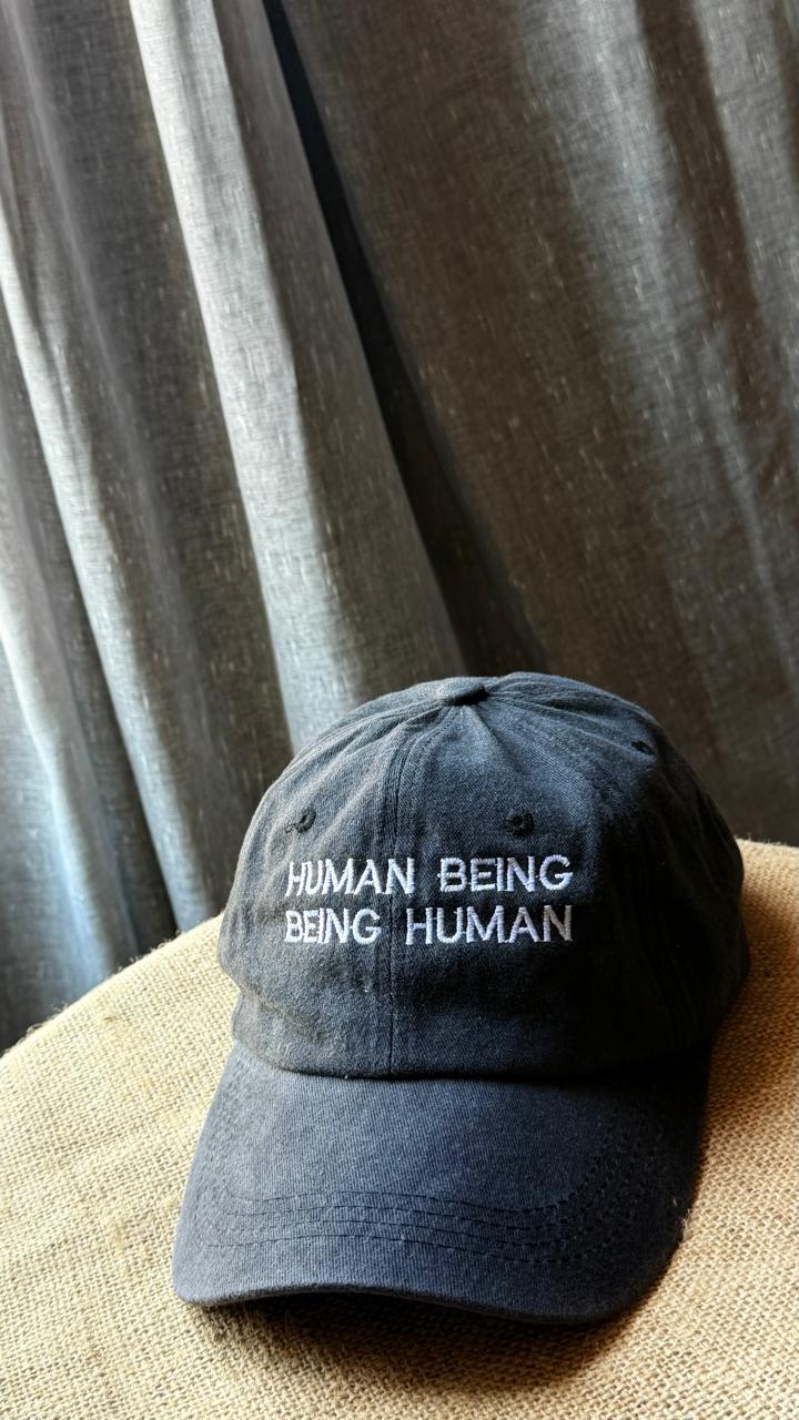 A dark navy baseball cap with 'HUMAN BEING BEING HUMAN' embroidered in white on the front, resting on a textured beige surface against a gray curtain background.